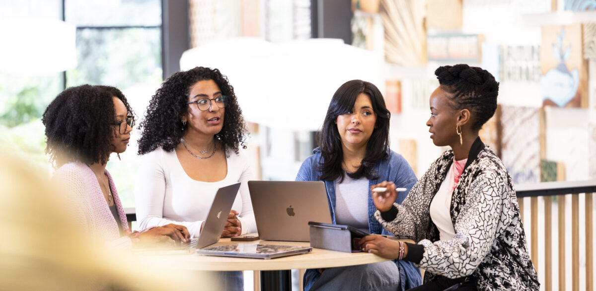 Four female students in discussion with each other while working on laptops and tablets