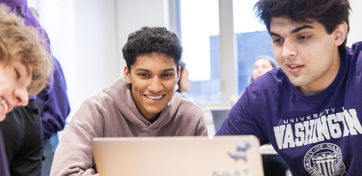 Three male students look at a laptop screen while seated at a table